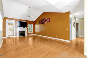 A spacious living room with light wood flooring, mustard-colored accent wall, ceiling fan, fireplace with a TV on top, large windows, and a door leading outside. Artwork hangs on the accent wall.