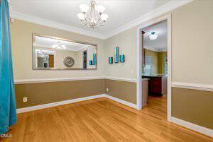 A dining room with light brown walls, white trim, hardwood floors, a large wall mirror, teal decorations, blue curtains, and a view into a kitchen with wooden cabinets. Two chandeliers hang from the ceiling.