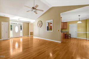 Spacious room with light wood flooring, vaulted ceiling, and ceiling fan. An entrance door with glass panels is visible, along with a kitchen area and breakfast bar in the background. Walls are painted a beige-yellow color.
