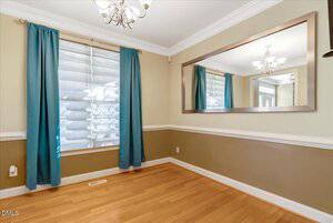 A room with light hardwood floors, beige walls with white trim, a large wall mirror, a window covered by white blinds and teal curtains, and a ceiling light fixture.