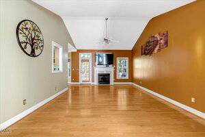 A spacious living room with light wood flooring, vaulted white ceiling, and earth-toned walls. A ceiling fan hangs above. The room features a fireplace with a TV above it and two windows, with wall art on both sides.