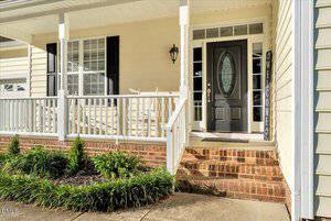 A front porch with white railings, brick steps, a black front door with a decorative glass oval, a welcome sign, two white rocking chairs, and trimmed green shrubs along the sidewalk.