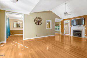 Spacious living room with light hardwood floors, high vaulted ceiling, ceiling fans, large windows, a fireplace with a TV above it, and an open doorway leading to a dining area. A tree wall art decorates one wall.