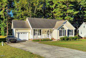 Single-story beige house with an attached garage, front porch with white railings, and large arched window, set on a spacious lawn with trees in the background and a black truck parked in the driveway.