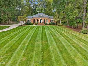 A large brick house with white trim sits at the end of a long, striped, freshly mowed lawn, surrounded by tall trees on both sides and a curved driveway on the left.