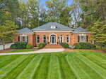 A one-story brick house with white trim and black shutters at 230 Saddletree Road, Oxford, featuring a central arched doorway, manicured lawn, neat shrubs, and trees in the background beneath a clear sky.