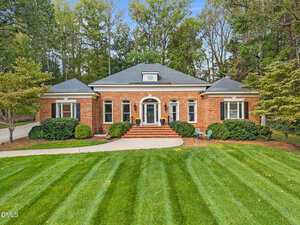 A one-story brick house with white trim and black shutters at 230 Saddletree Road, Oxford, featuring a central arched doorway, manicured lawn, neat shrubs, and trees in the background beneath a clear sky.