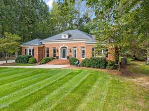Single-story red brick house with black shutters, a gray roof, and a central entrance. The lawn is well-maintained with green grass, shrubs, and mature trees surrounding the property. Steps lead to the front door.