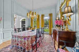 A formal dining room with a long table covered by a patterned cloth, dark chairs, a gold chandelier, a sideboard with lamps and flowers, yellow curtains, and a large window overlooking a garden.
