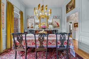 A dining room features a long table with a floral tablecloth, six dark wooden chairs, a gold chandelier, and a sideboard with lamps and flowers. Artwork and decorative plates hang on pale blue paneled walls.