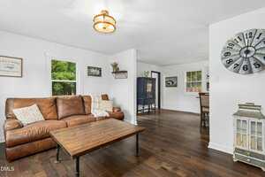 A living room with a brown leather sofa, wooden coffee table, white walls, dark wood floors, and a large wall clock. A window shows greenery outside, and a dining area is visible in the background.