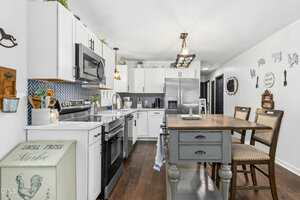 Modern kitchen with white cabinets, stainless steel appliances, blue tile backsplash, and a wooden dining table with four chairs. Decorative accents and kitchen items are displayed on counters and walls.