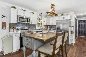 A modern kitchen features white cabinets, stainless steel appliances, a central island with two chairs, a patterned blue backsplash, wooden floors, and decorative plants on top of the cabinets.