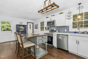 Bright kitchen with white cabinets, stainless steel appliances, a patterned blue backsplash, large windows, a wooden dining table with two chairs, light fixture above, and hardwood floors. Door and hutch visible in the background.