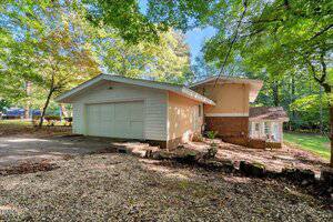 A two-story house with light siding and brick accents is surrounded by trees. The house features a large attached garage, a gravel driveway, and a glass-enclosed porch area on the right. The yard is shaded with mature trees.