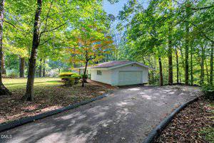 A single-story white house with a two-car garage sits among tall trees. A curved driveway leads to the garage, and sunlight filters through the leaves, casting shadows on the ground.