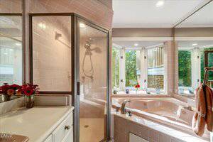 A bathroom with pink tile walls, a glass shower enclosure, a pink corner bathtub with jets, a white countertop with a vase of red flowers, and large windows letting in natural light.