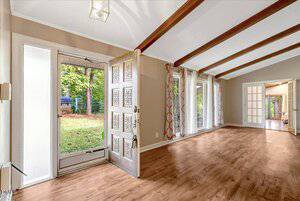 A spacious living room with wood flooring, exposed ceiling beams, large windows with patterned curtains, and a partially open front door leading to a grassy backyard. Double doors are visible at the far end of the room.