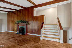A living room with wood-paneled walls, a fireplace with greenery on the mantel, wood flooring, and a split staircase leading to two different levels. The area is well-lit with neutral tones.