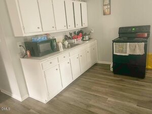 A kitchen with white cabinets, a black microwave, various kitchen items on the counter, a black stove with pots and towels, and wood-look vinyl flooring. A framed sign hangs on the light-colored wall above the stove.