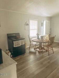 A small kitchen area with a black stove, a wooden dining table with three chairs, and two windows letting in natural light. The floor is wood-look laminate, and the walls are light-colored.