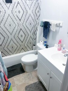 A small bathroom with a white sink and cabinet, toilet, and bathtub with a gray patterned shower curtain. Towels hang on a rack, and toiletries are on the sink. There are gray bath mats on the floor.
