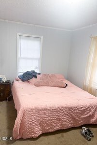 A bedroom with light-colored walls, a bed covered in a pink quilt with pillows and a blanket, a small wooden nightstand with items on it, and a pair of shoes on the carpeted floor. A window is behind the bed.