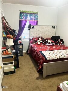 A small bedroom with a bed covered in red and white bedding, assorted pillows, boxes stacked beside the bed, bags hanging on the wall, and a window with a purple curtain and a decorative valance.