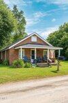 A small brick house at 1300 Railroad Street features a covered front porch with blue chairs, a mailbox on a wooden post, and a grassy lawn bordered by trees under a partly cloudy sky.