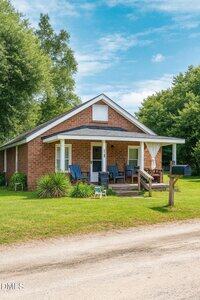 A small brick house at 1300 Railroad Street features a covered front porch with blue chairs, a mailbox on a wooden post, and a grassy lawn bordered by trees under a partly cloudy sky.