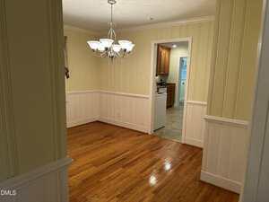 A dining room with wood floors, cream-colored walls with white wainscoting, and a ceiling light fixture. An open doorway leads to a kitchen with tile flooring and wooden cabinets.