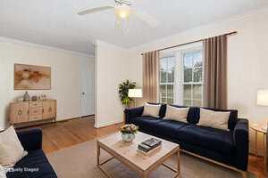A living room with a dark blue sofa, a wooden coffee table, a sideboard with wall art above it, a potted plant, two table lamps, beige curtains, hardwood floors, and a ceiling fan with a light.
