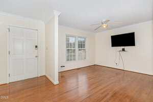 Empty living room with light-colored walls, hardwood floors, a ceiling fan, a wall-mounted TV, and large windows letting in natural light. The entrance door is visible on the left side of the image.
