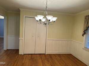 A dining room with light-colored wood paneling, hardwood floors, a white double-door closet, a silver chandelier with white shades, and a window with a partial valance on the right.