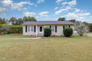 A small, single-story beige house with black shutters and a dark roof sits on a grassy lawn. A concrete walkway leads to the front door, and trimmed bushes are planted near the entrance. Trees and blue sky with clouds are in the background.