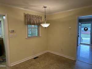 A small dining area with pale yellow walls, a window with a curtain, a hanging light fixture, and tile flooring; to the right is a doorway leading to a front entry with wood flooring, visible outdoors.