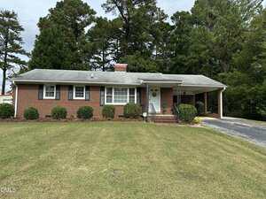 A single-story brick house at 1224 Oakridge Avenue features a gray roof, white-framed windows, and a carport on the right. The grassy front lawn is accented by trimmed bushes and tall trees in the background.