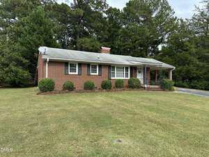 Single-story brick house with black shutters, a gray shingled roof, a small covered porch, and a red brick chimney. The home is surrounded by bushes, a large lawn, and tall trees in the background.