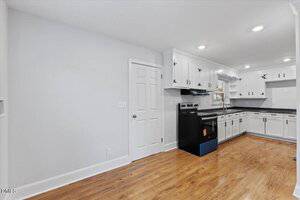 A kitchen with white cabinets, black appliances, and wood flooring. The walls are light-colored, and there is a door on the left side. Recessed ceiling lights provide illumination. The space appears clean and empty.