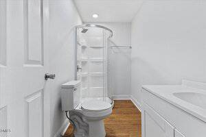 A small bathroom with a white toilet, a corner glass shower with built-in shelves, a white vanity with sink, and wood flooring. The walls and ceiling are white, and there is recessed lighting.