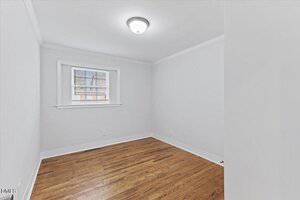 A small, empty room with white walls, a ceiling light fixture, a single window with blinds, and wooden flooring. The room is well-lit and unfurnished.