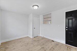A small room with white walls, light wood flooring, a closed white door, a black door, a window with blinds, baseboards, and a ceiling-mounted light fixture.