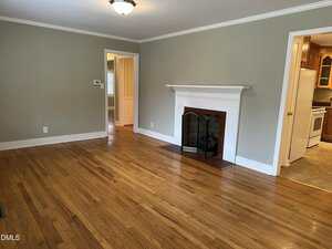 A living room with hardwood floors, a white fireplace with a screen, light green walls, white trim, and doorways leading to a hallway and a kitchen with wooden cabinets. Ceiling light and thermostat visible.