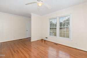 An empty room with light beige walls, hardwood flooring, a white ceiling fan, crown molding, a large window with blinds, and a closed white door on the left wall.
