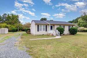 Single-story beige house with black shutters, a small front porch, and white railings. Gravel driveway curves toward the house. Green lawn, bushes, trees, and a small shed are visible in the background under a partly cloudy sky.