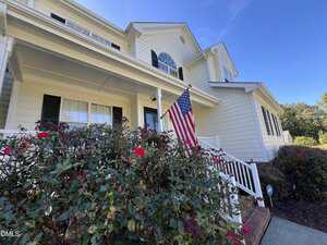 Two-story white house with black shutters, a front porch, and an American flag. Red rose bushes line the walkway, and the sky is clear and blue.