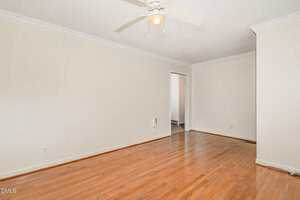 A room with light-colored walls and crown molding, a white ceiling fan with light, hardwood floors, and an open doorway leading to another space. No furniture or decor is present.