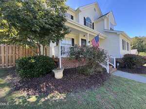 Two-story beige house with white trim, a covered front porch, and brick steps. An American flag hangs by the entrance. There are bushes, a small tree, and a planter in the front yard, with a wooden fence on one side.