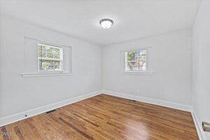 Empty room with hardwood floors, white walls, two small windows with white blinds, a ceiling light fixture, and baseboard trim. Natural light comes through the windows.