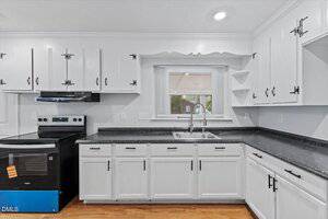 A kitchen with white cabinets, black countertops, an electric stove with a black range hood, a stainless steel sink under a window with blinds, and hardwood floors.